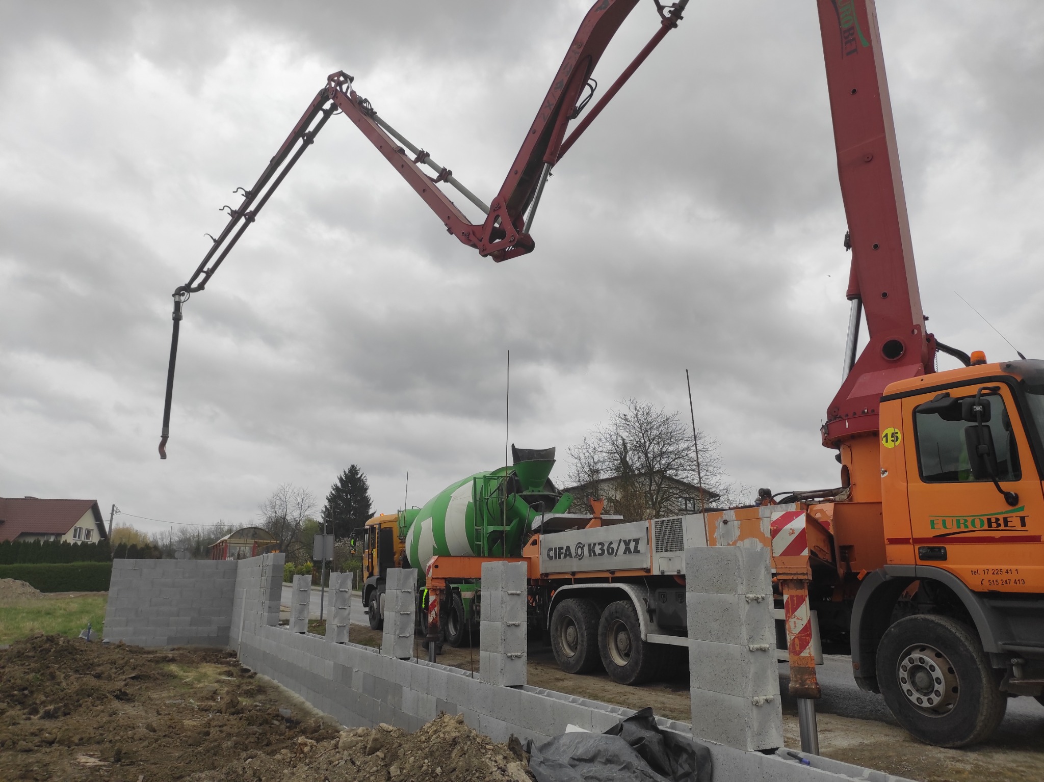 Fundamenty budynku w trakcie budowy z użyciem betonomieszarki CIFA K36/XZ i pompy do betonu Eurobet na tle pochmurnego nieba.