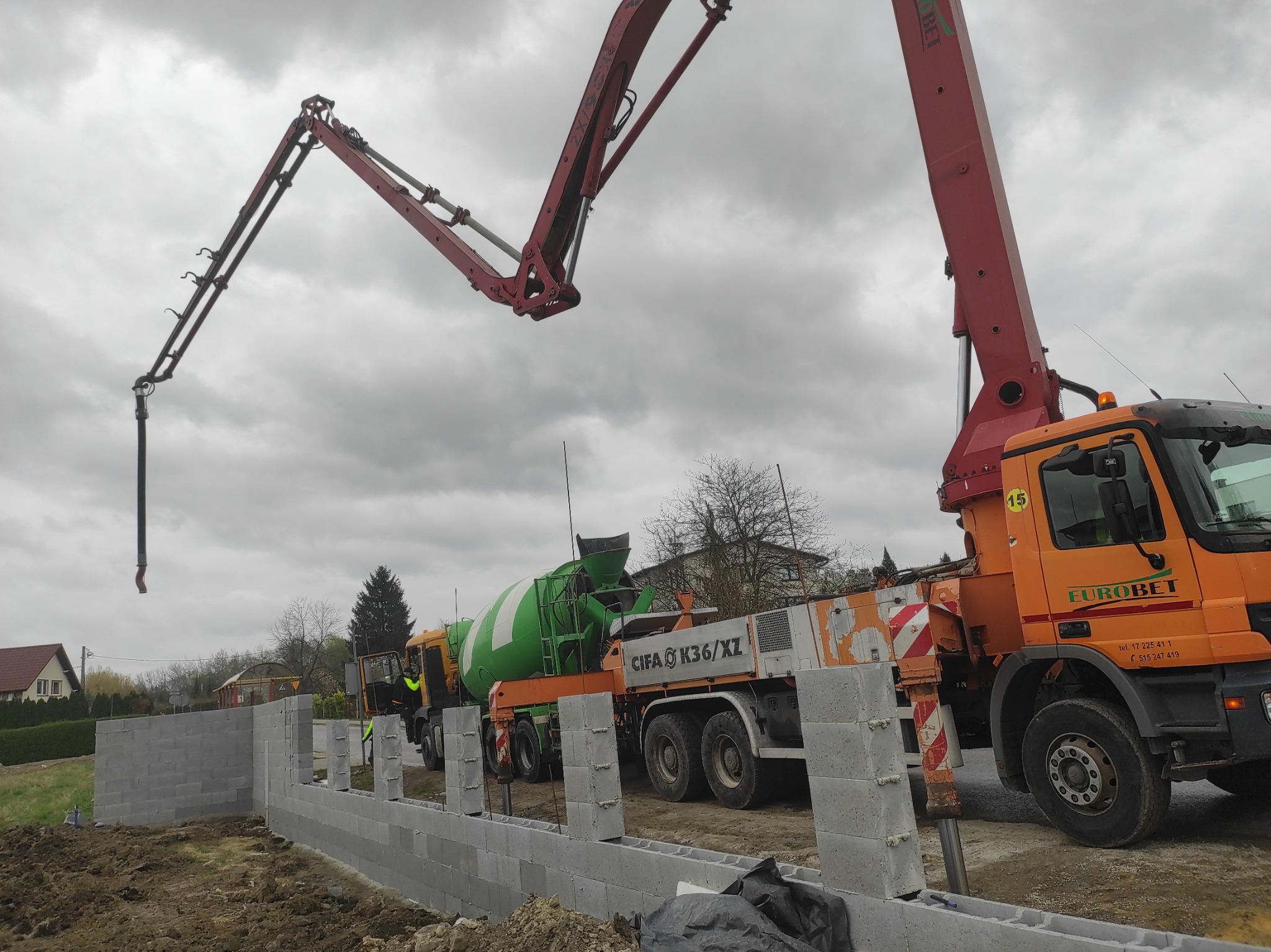 Budowa fundamentów: pompa do betonu Eurobet w akcji, w tle zielona betoniarka CIFA K36/XZ, mur z szarych bloczków betonowych w trakcie wznoszenia.