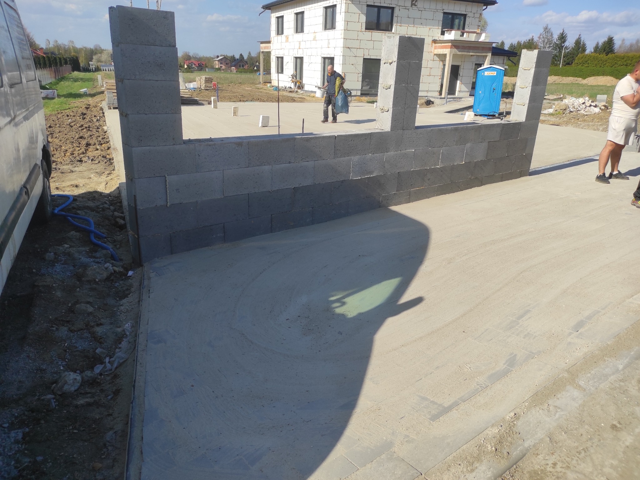 Wznoszenie muru z szarego bloczka betonowego na fundamencie. W tle budynek w trakcie budowy i przenośna toaleta.