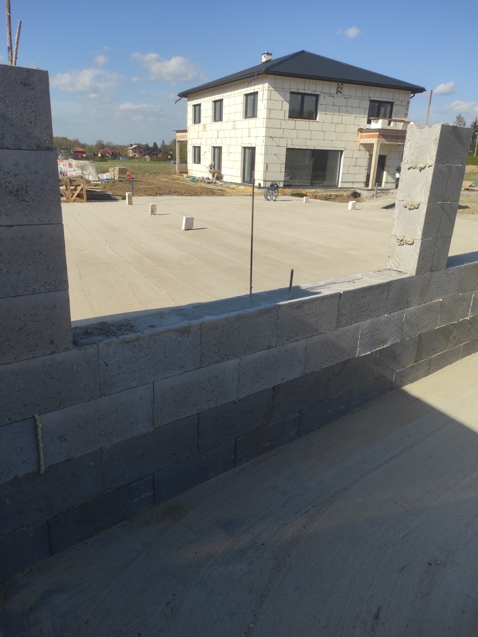 Wznoszenie ścian fundamentowych z bloczków betonowych, widoczny stan surowy otwarty budynku w tle, teren budowy, słoneczny dzień.