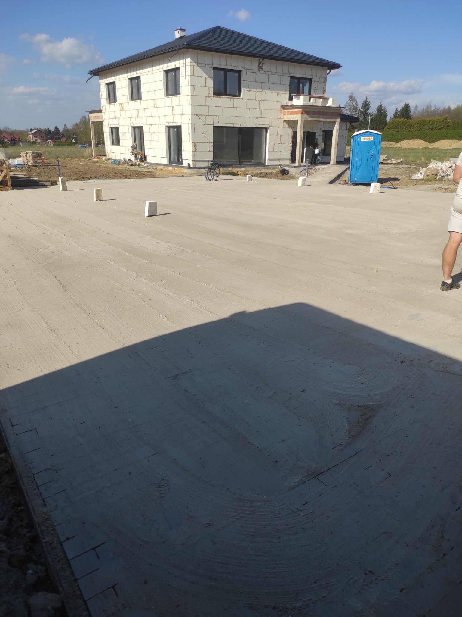 Przygotowany teren pod budowę domu jednorodzinnego. Widoczna ubita powierzchnia, białe bloczki, częściowo wykończony budynek i przenośna toaleta. Słoneczny dzień.