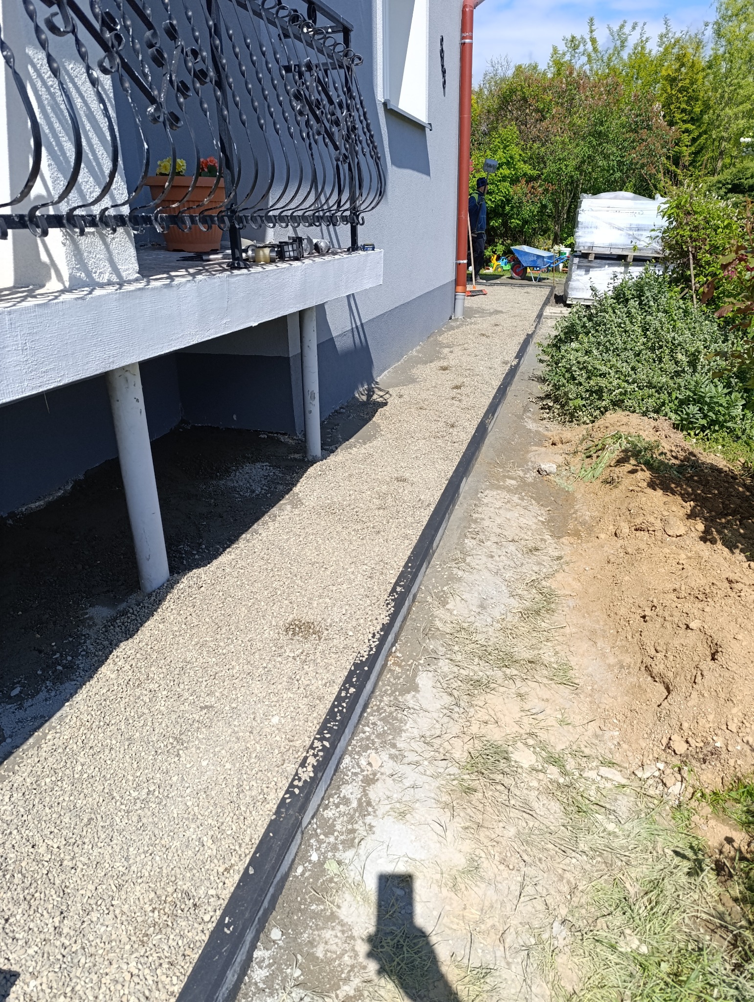 Przygotowanie podłoża z kruszywa pod kostkę brukową wzdłuż budynku z balkonem. Widoczne obrzeża i wyrównany teren. W tle materiały budowlane i pracownik.