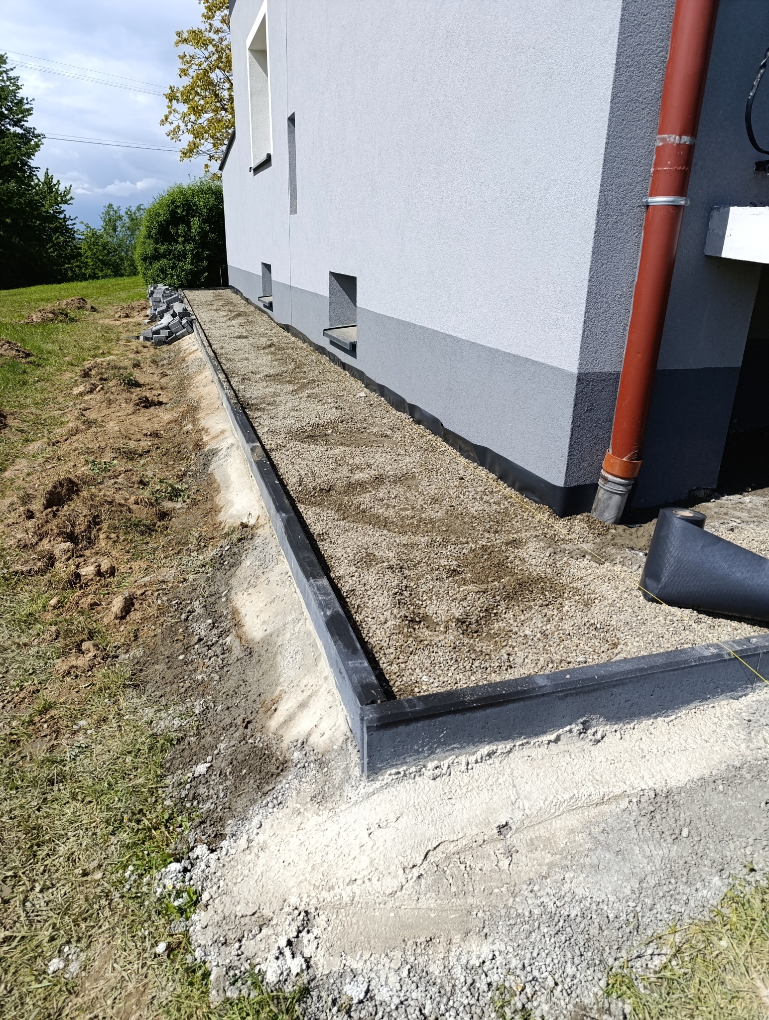 Przygotowanie podłoża pod kostkę brukową przy budynku: obrzeża zamontowane, podsypka z kruszywa wysypana, widoczne rolki geowłókniny.