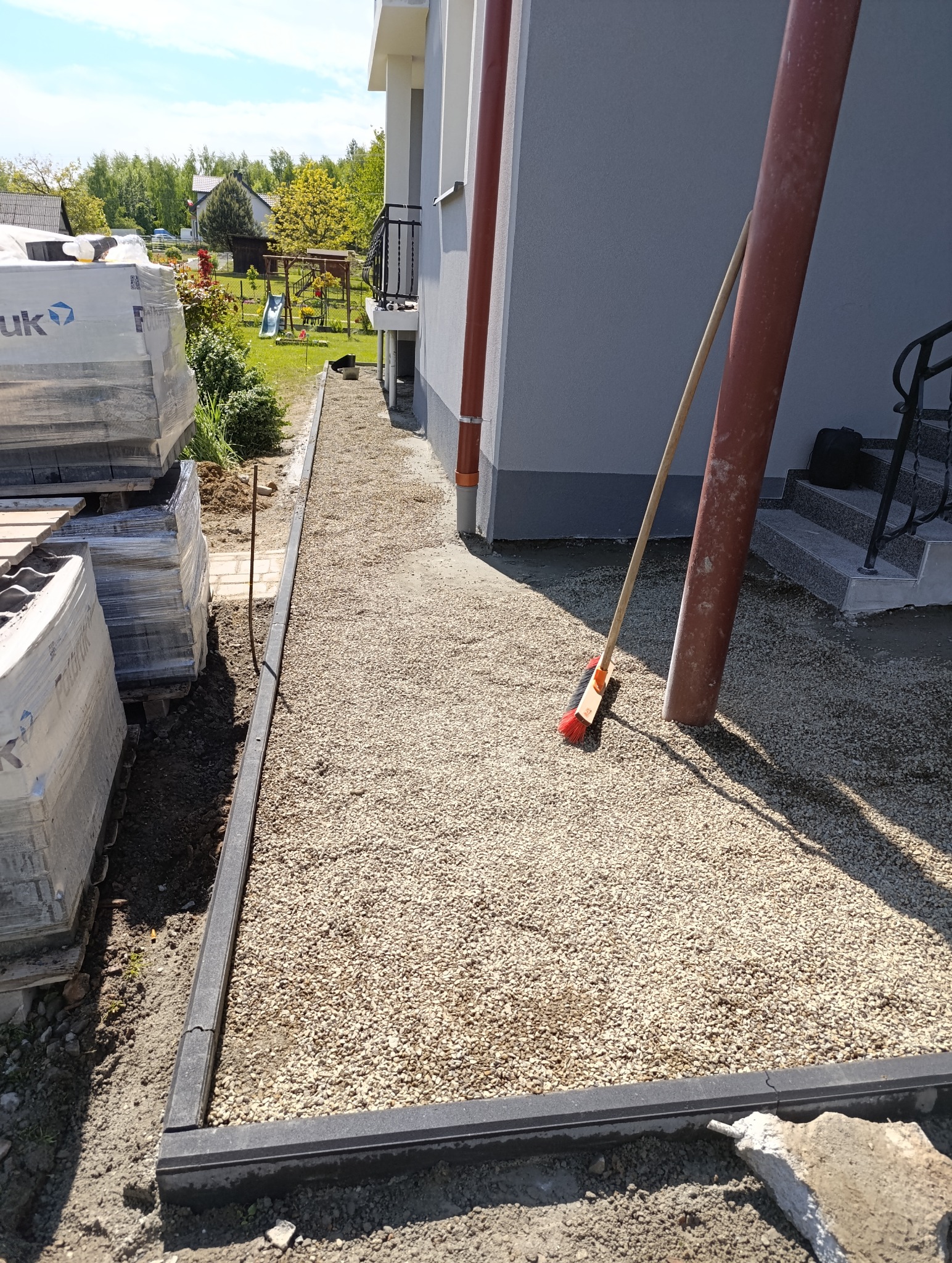 Przygotowanie podłoża pod ścieżkę z kostki: obrzeża, podsypka żwirowa, palety kostki brukowej obok. Widok z góry, w tle budynek i zieleń.