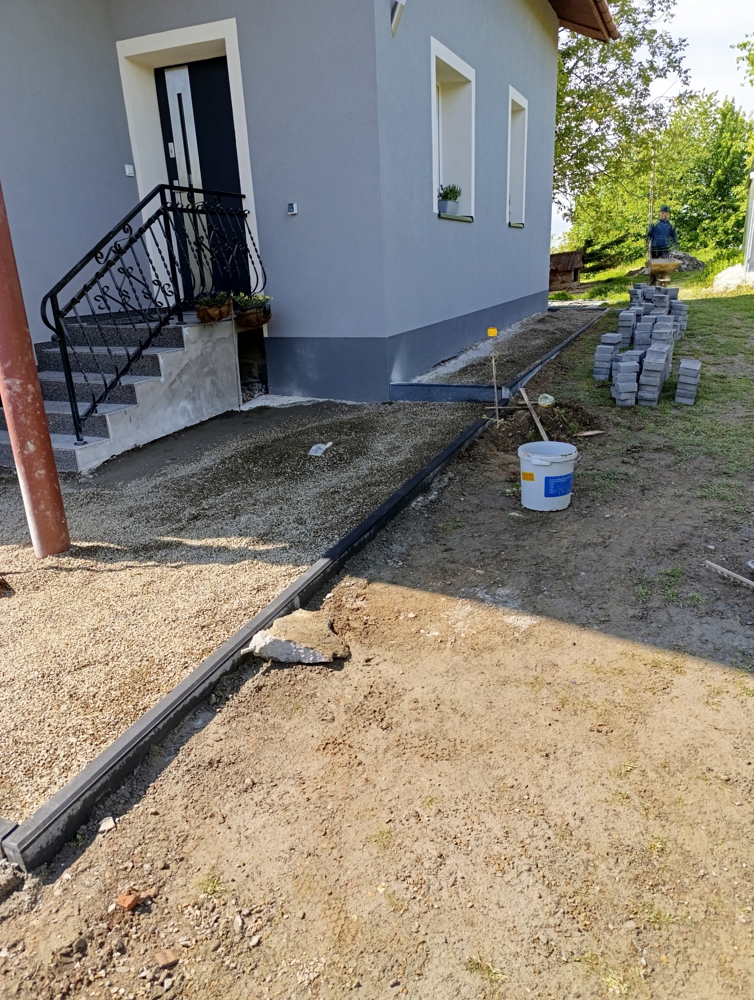 Przygotowanie terenu pod kostkę brukową: ułożone obrzeża, podsypka z kruszywa, kostka w tle, dom z schodami i roślinami w donicach. Prace ziemne w toku.