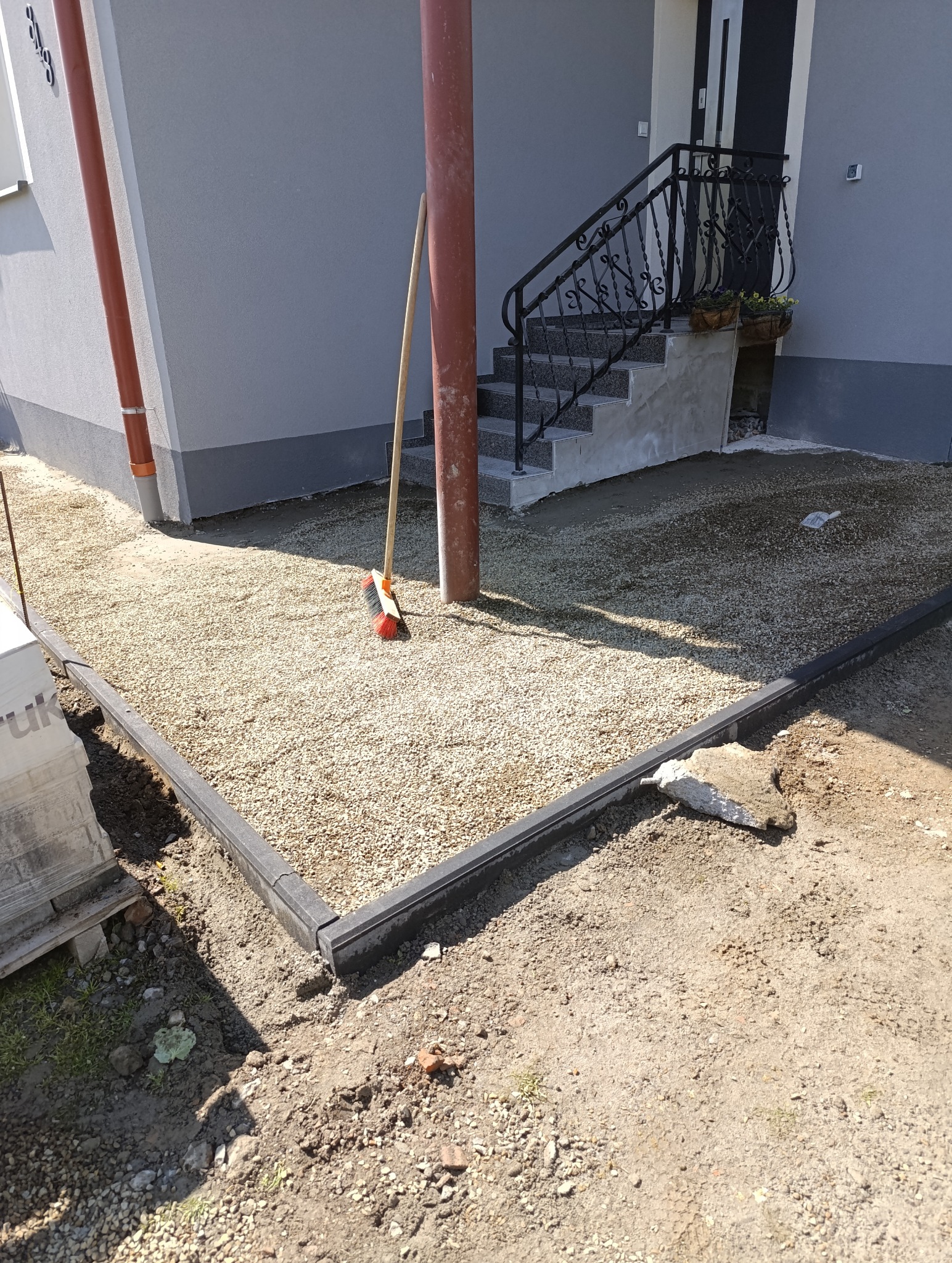 Przygotowany teren pod kostkę: podsypka żwirowa z obrzeżami, widoczne schody wejściowe do domu, grabie oparte o rynnę. Etap prac brukarskich.