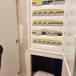 Dawid Górnicki - Otwarta skrzynka elektryczna z widocznymi bezpiecznikami i okablowaniem, zlokalizowana w ścianie z widocznymi śladami po remoncie; widoczne dokumenty i przewody w dolnej części szafki.