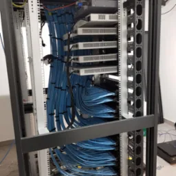 Widok z bliska na szafę serwerową z zamontowanymi urządzeniami i uporządkowanymi niebieskimi kablami Ethernet, widoczne metalowe szyny montażowe i część podłogi.