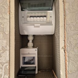 Dawid Górnicki - Dwa panele elektryczne Schneider zamontowane w ścianie z izolacją i tapetą w kwiaty, widoczne bezpieczniki B10 i B16.