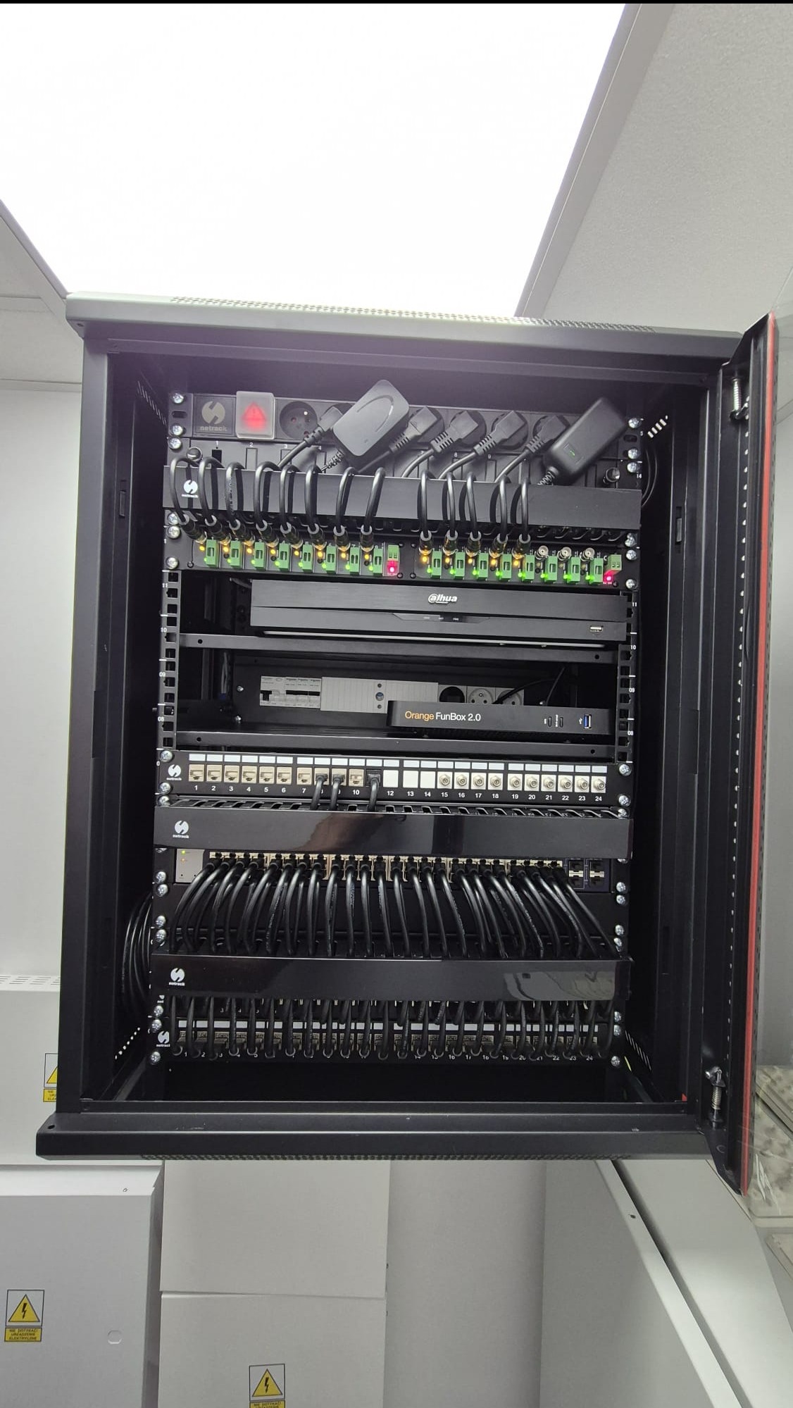 Otwarta szafa rack z zainstalowanym sprzętem sieciowym, widoczne patch panele, zasilacze, rejestrator Dahua i router Orange FunBox 2.0, kable uporządkowane w grzebieniach.