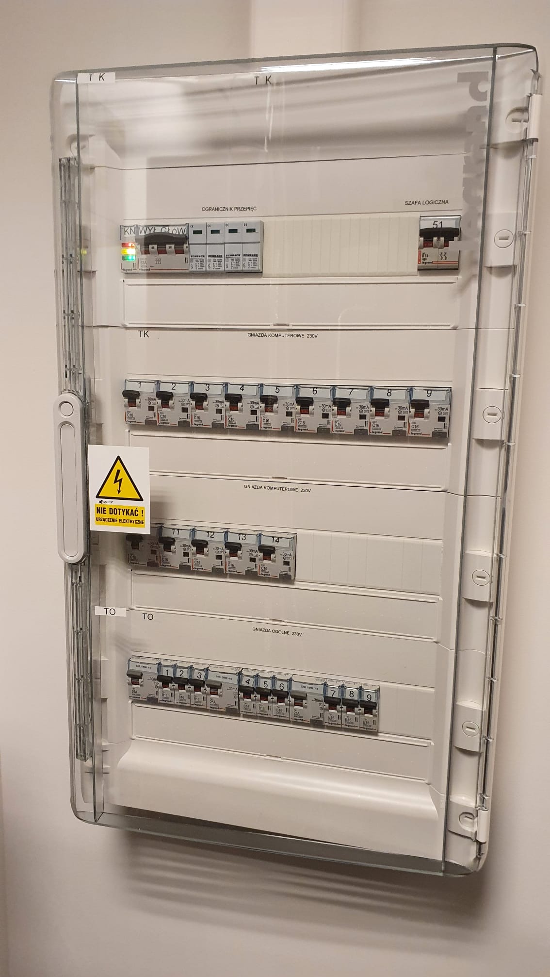 Rozdzielnica elektryczna z przezroczystą obudową, zawierająca wyłączniki nadprądowe i różnicowoprądowe, z oznaczeniami 'Gniazda komputerowe 230V' oraz naklejką ostrzegawczą 'Nie dotykać! Urządzenie...