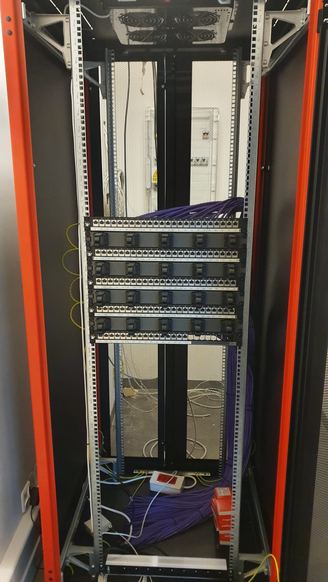 Widok z przodu na częściowo wypełnioną szafę serwerową z patch panelem, fioletowymi kablami i pomarańczową ramą, widoczna biała listwa zasilająca i opakowania po kablach.