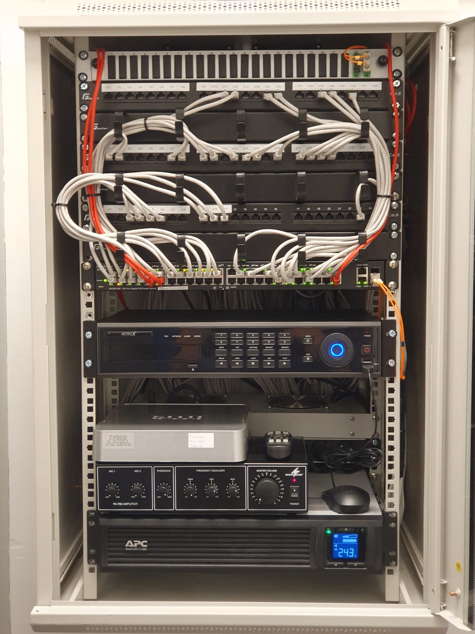 Widok z bliska na szafę rack z zainstalowanymi urządzeniami sieciowymi, patch panele z podłączonymi kablami Ethernet, rejestrator INTROX z niebieskim podświetleniem, wzmacniacz PA-702 oraz zasilacz...