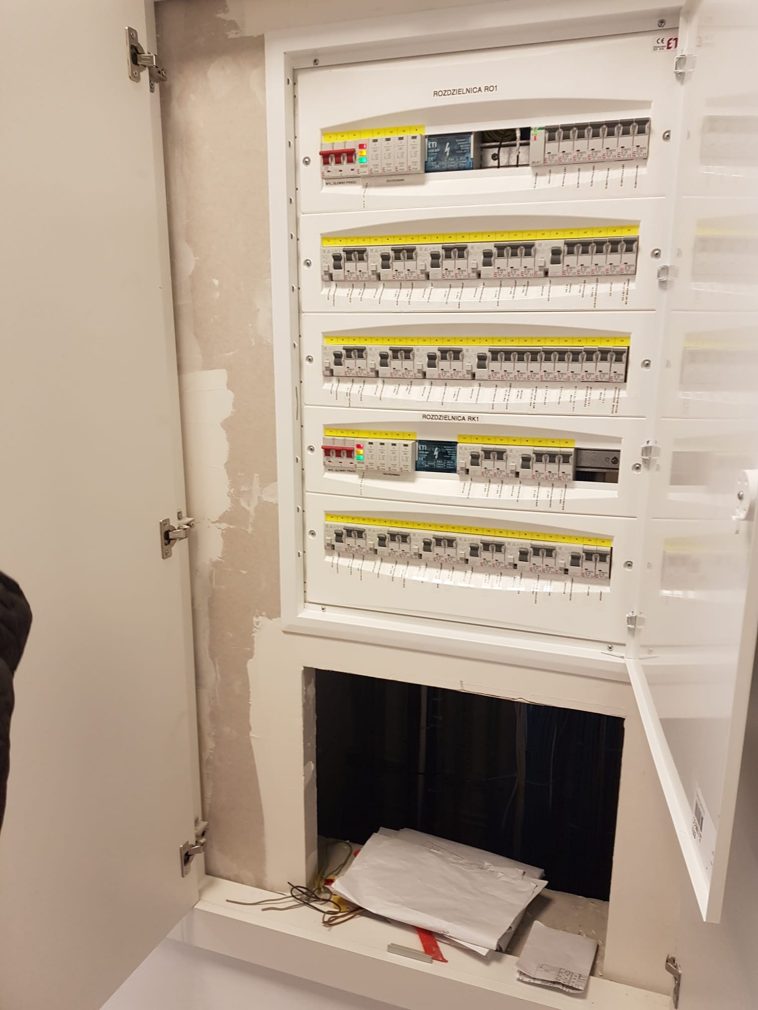 Otwarta skrzynka elektryczna z widocznymi bezpiecznikami i okablowaniem, zlokalizowana w ścianie z widocznymi śladami po remoncie; widoczne dokumenty i przewody w dolnej części szafki.