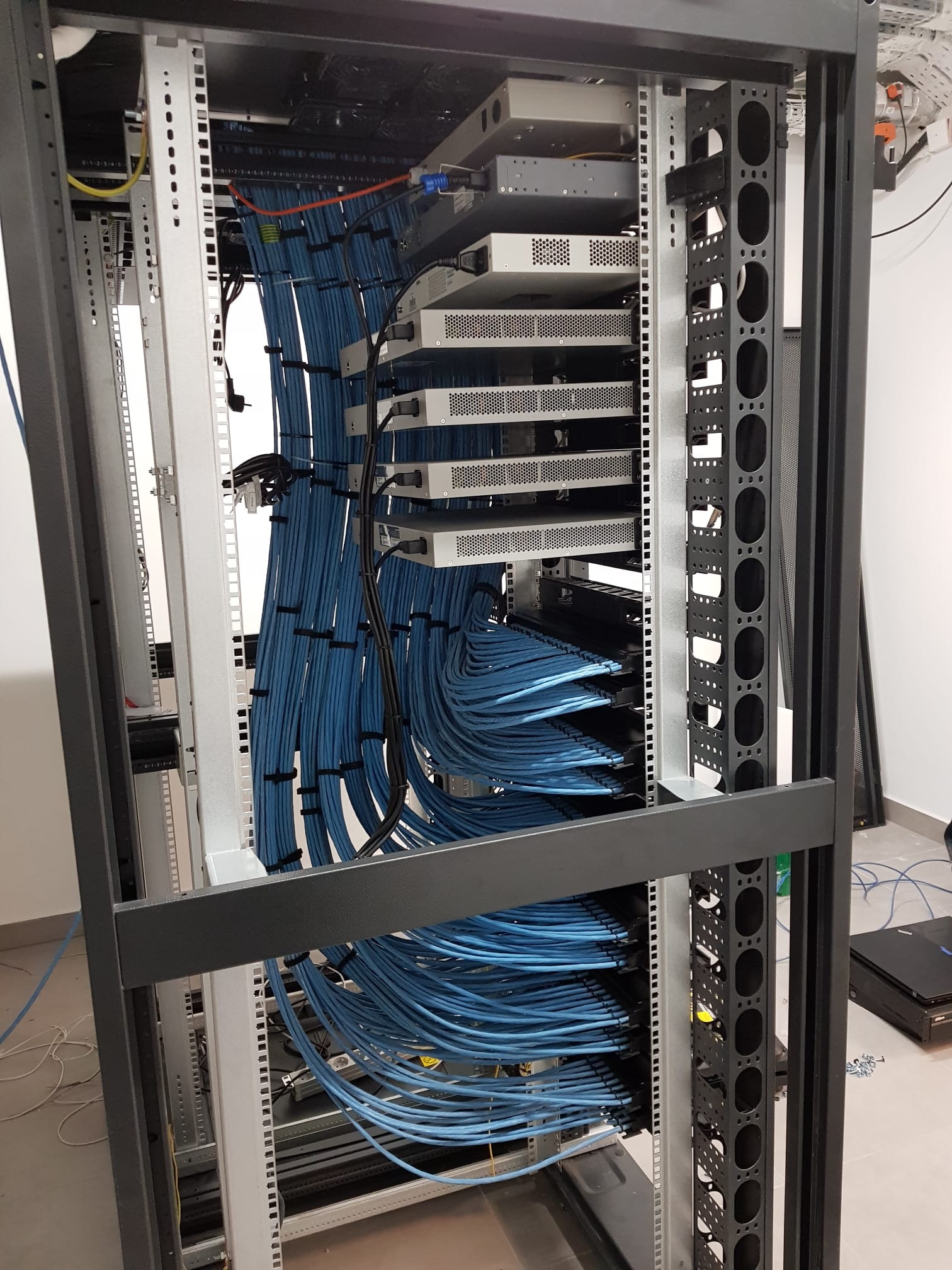 Widok z bliska na szafę serwerową z zamontowanymi urządzeniami i uporządkowanymi niebieskimi kablami Ethernet, widoczne metalowe szyny montażowe i część podłogi.