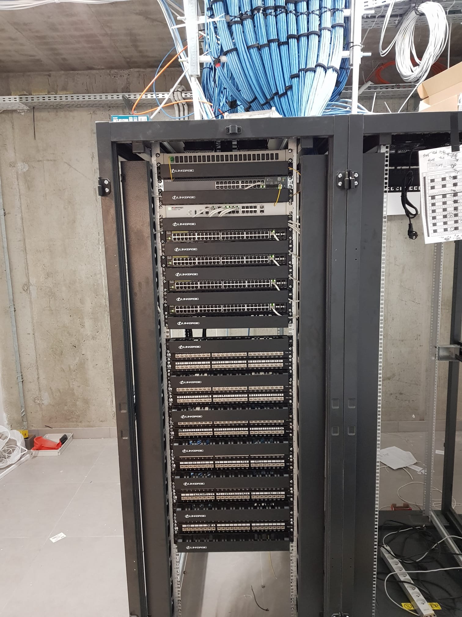 Szafa rack z zamontowanymi przełącznikami sieciowymi marki Linkbasic i Fortinet, okablowanie w kolorze niebieskim u góry, betonowa ściana w tle.