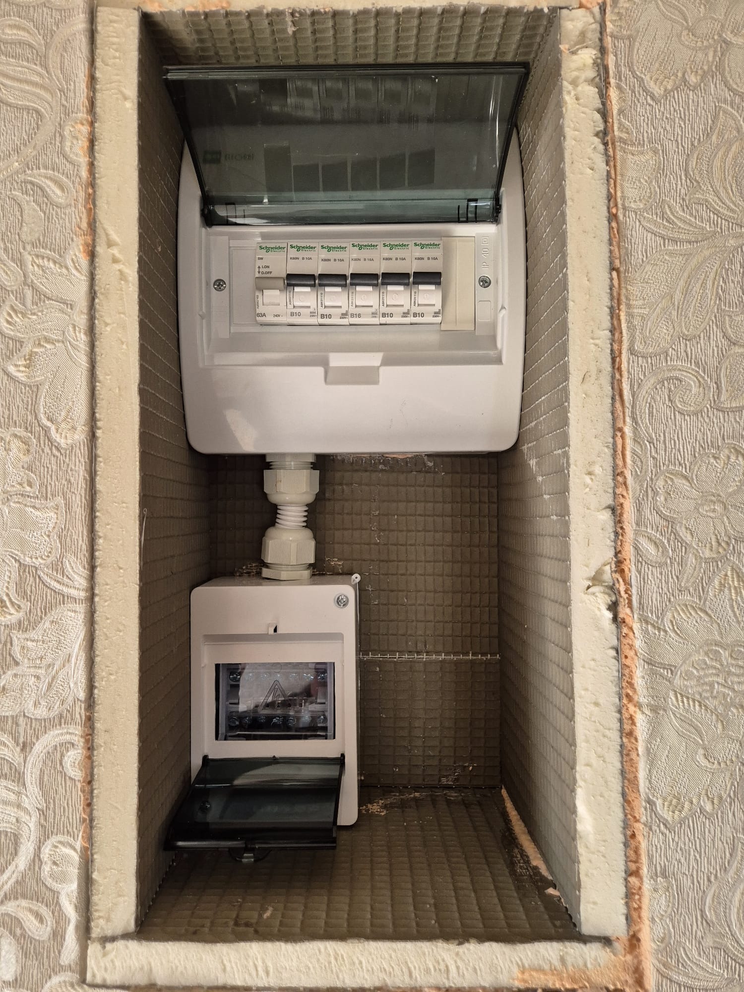 Dwa panele elektryczne Schneider zamontowane w ścianie z izolacją i tapetą w kwiaty, widoczne bezpieczniki B10 i B16.