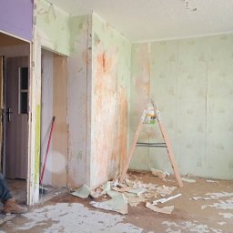 Małgorzata Grzesik MAL - BUD - Przygotowanie do malowania ścian w Chorzowie: Ślady po zerwanym starym wystroju, drabina malarska i pozostałości tapet na podłodze. Fragment widocznego pomieszczenia.