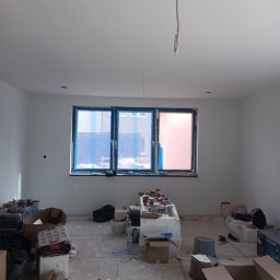 Małgorzata Grzesik MAL - BUD - Pokój w trakcie remontu: ściany po gładzi, zabezpieczone okna, materiały budowlane na podłodze. Widoczne przewody wystające z sufitu.