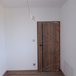 Małgorzata Grzesik MAL - BUD - Pokój po remoncie: drewniana podłoga, świeżo pomalowane białe ściany, zabezpieczone folią drzwi z drewnianą okleiną i czarnym paskiem, wystający kabel z sufitu.