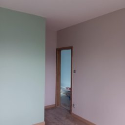 Małgorzata Grzesik MAL - BUD - Pomalowane na dwa kolory wnętrze z drewnianą podłogą i listwami przypodłogowymi. W tle widoczne otwarte drzwi do innego pomieszczenia z niebieską ścianą.