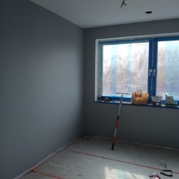 Małgorzata Grzesik MAL - BUD - Pomieszczenie po malowaniu ścian na szaro, z widocznymi narzędziami malarskimi przy oknie i na podłodze. Okno zabezpieczone folią. Wystający kabel z sufitu.