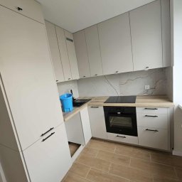 Małgorzata Grzesik MAL - BUD - Jasna, nowoczesna kuchnia z drewnianą podłogą i marmurowym backsplashiem. Niebieskie wiadro na blacie dodaje koloru. Minimalistyczne uchwyty szafek.