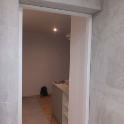 Małgorzata Grzesik MAL - BUD - Wykończone przejście w łazience z szarymi płytkami, widok na wnętrze z szafką i torbą. Delikatne oświetlenie sufitowe. Minimalistyczny design.