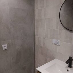 Małgorzata Grzesik MAL - BUD - Wykończona łazienka z szarymi płytkami imitującymi kamień, okrągłym lustrem i umywalką nablatową z drewnianą szafką. Widoczne gniazdka elektryczne.