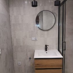 Małgorzata Grzesik MAL - BUD - Wykończona łazienka z szarą, pionową glazurą, okrągłym lustrem, drewnianą szafką pod umywalką i czarnymi lampami wiszącymi.