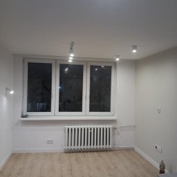 Małgorzata Grzesik MAL - BUD - Jasne wnętrze po remoncie: panele podłogowe, białe ściany, okno z grzejnikiem. Minimalistyczne oświetlenie punktowe, gniazdka elektryczne i butelka w rogu.