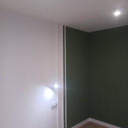 Małgorzata Grzesik MAL - BUD - Pomalowane na biało i oliwkowo ściany w rogu pokoju, z widoczną listwą przypodłogową i włącznikiem światła, oświetlone punktowym światłem.