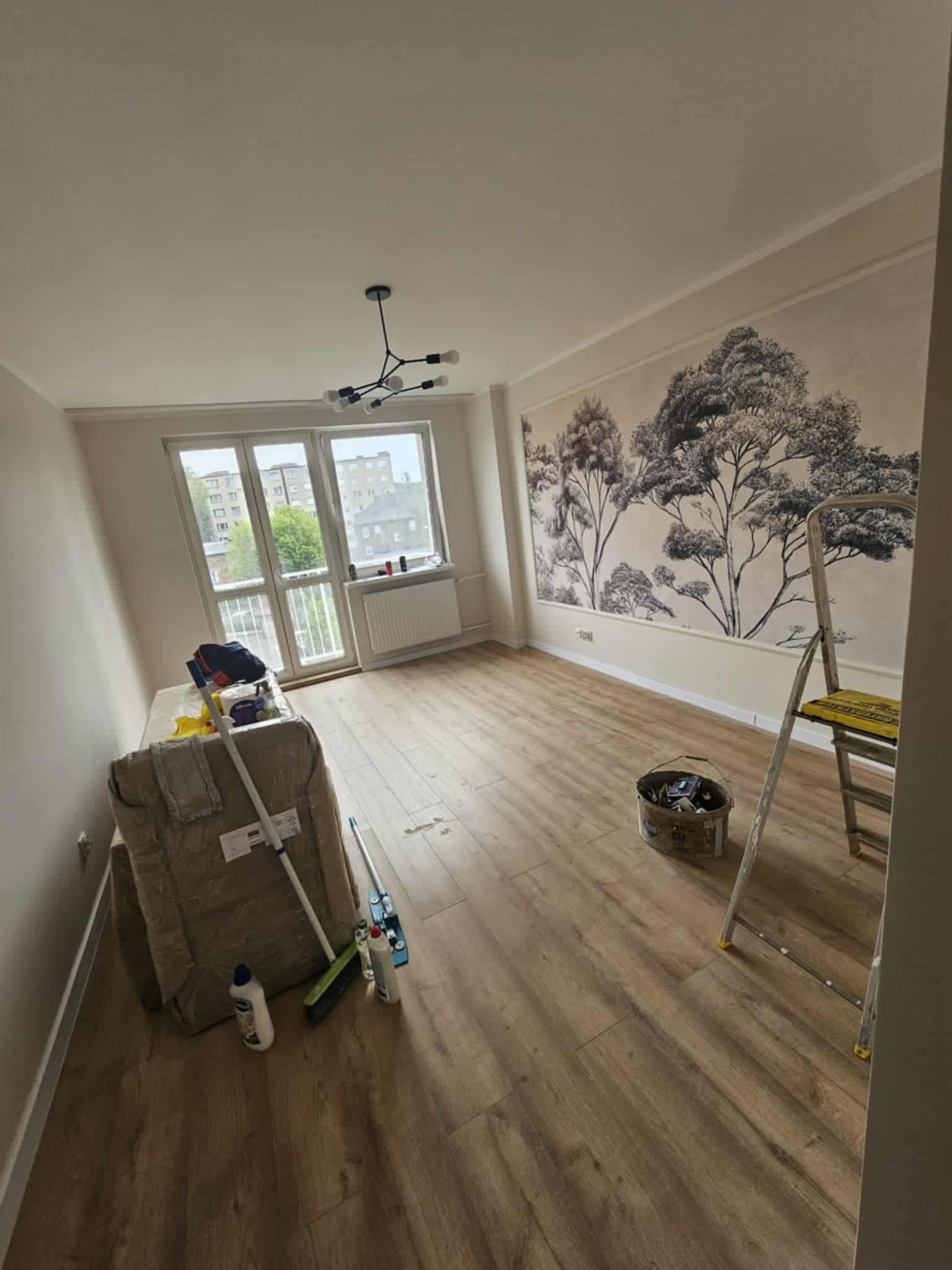 Wykończone wnętrze pokoju z panelami, na ścianie fototapeta z motywem drzew, drabina malarska, wiadro z farbą i akcesoria malarskie. Nowoczesna lampa sufitowa.