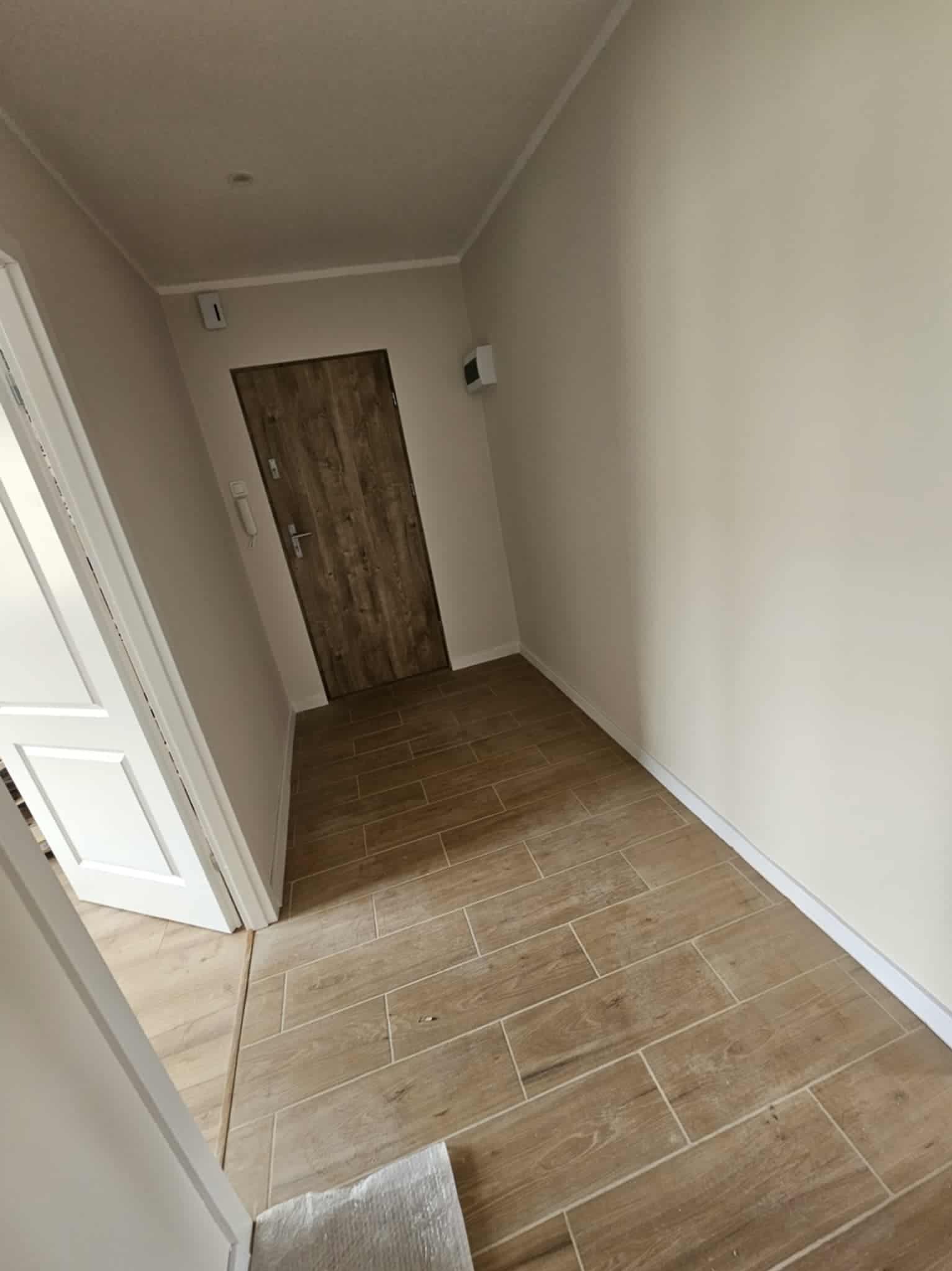 Korytarz po remoncie: beżowe ściany, drewnopodobna podłoga z płytek, biała listwa przypodłogowa i futryna. Drzwi wejściowe w odcieniach brązu.