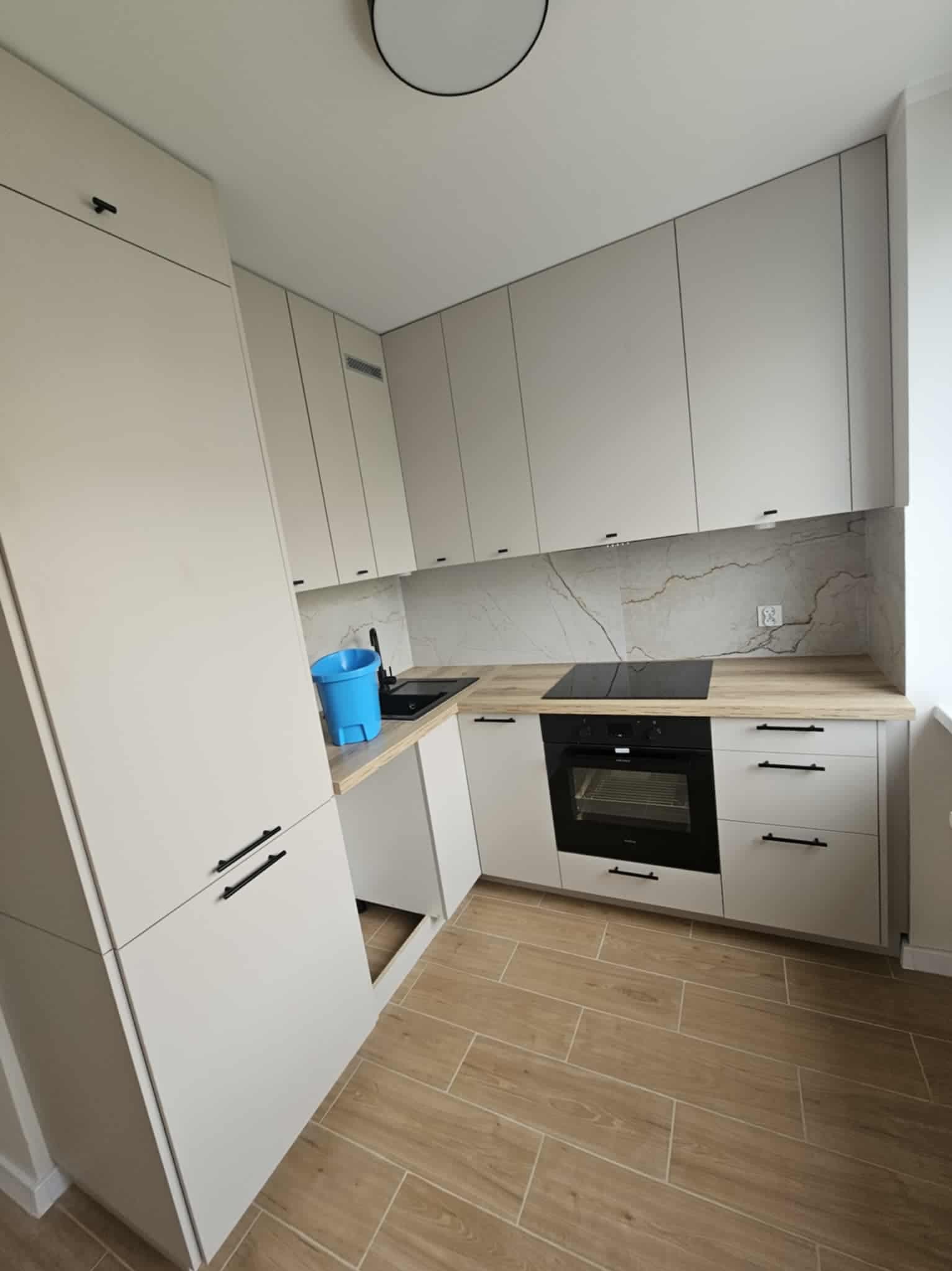 Jasna, nowoczesna kuchnia z drewnianą podłogą i marmurowym backsplashiem. Niebieskie wiadro na blacie dodaje koloru. Minimalistyczne uchwyty szafek.