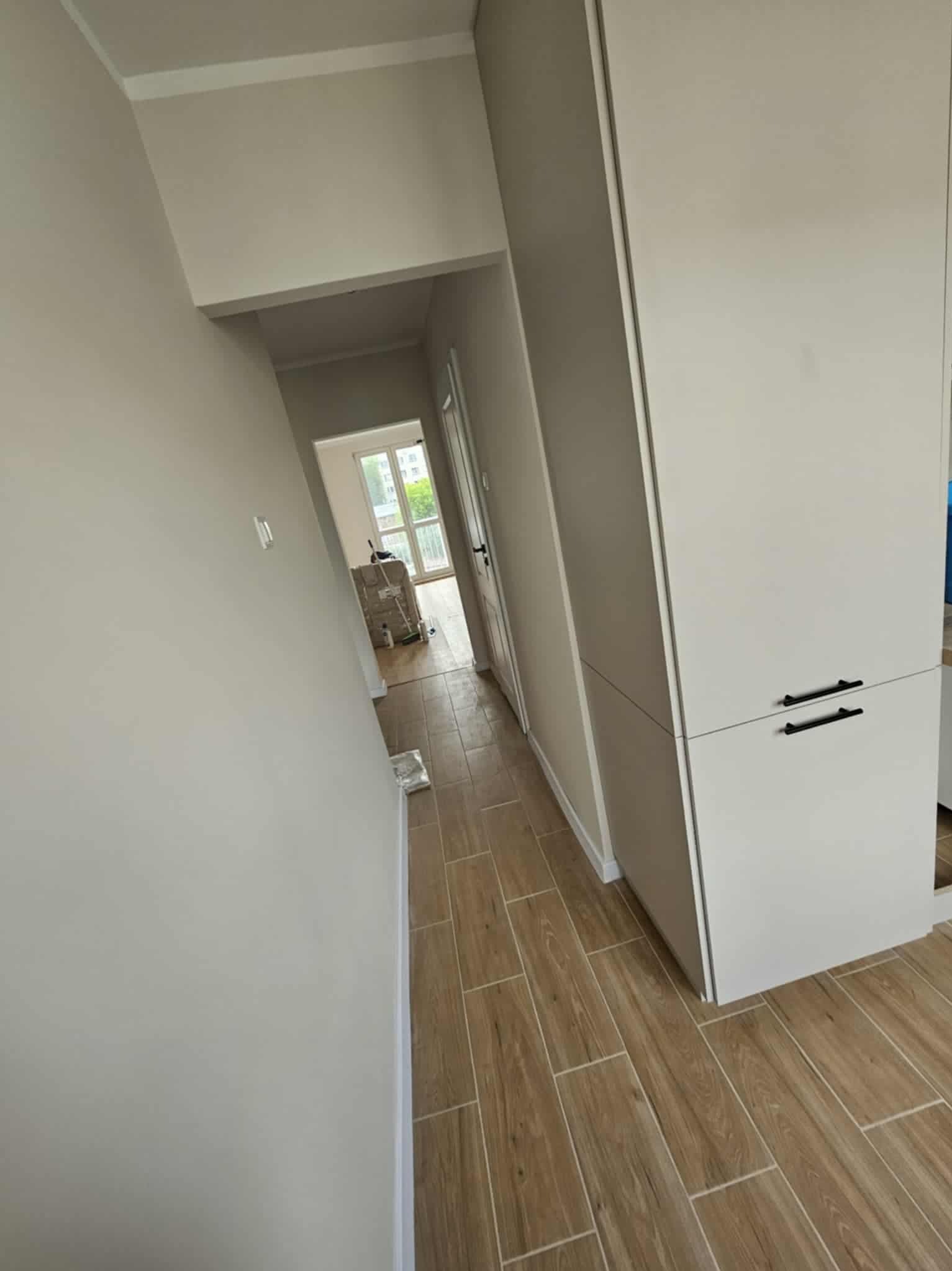 Jasny korytarz po remoncie: kremowe ściany, drewnopodobna terakota, biała zabudowa meblowa z czarnymi uchwytami. Widok na jasne pomieszczenie z oknem.