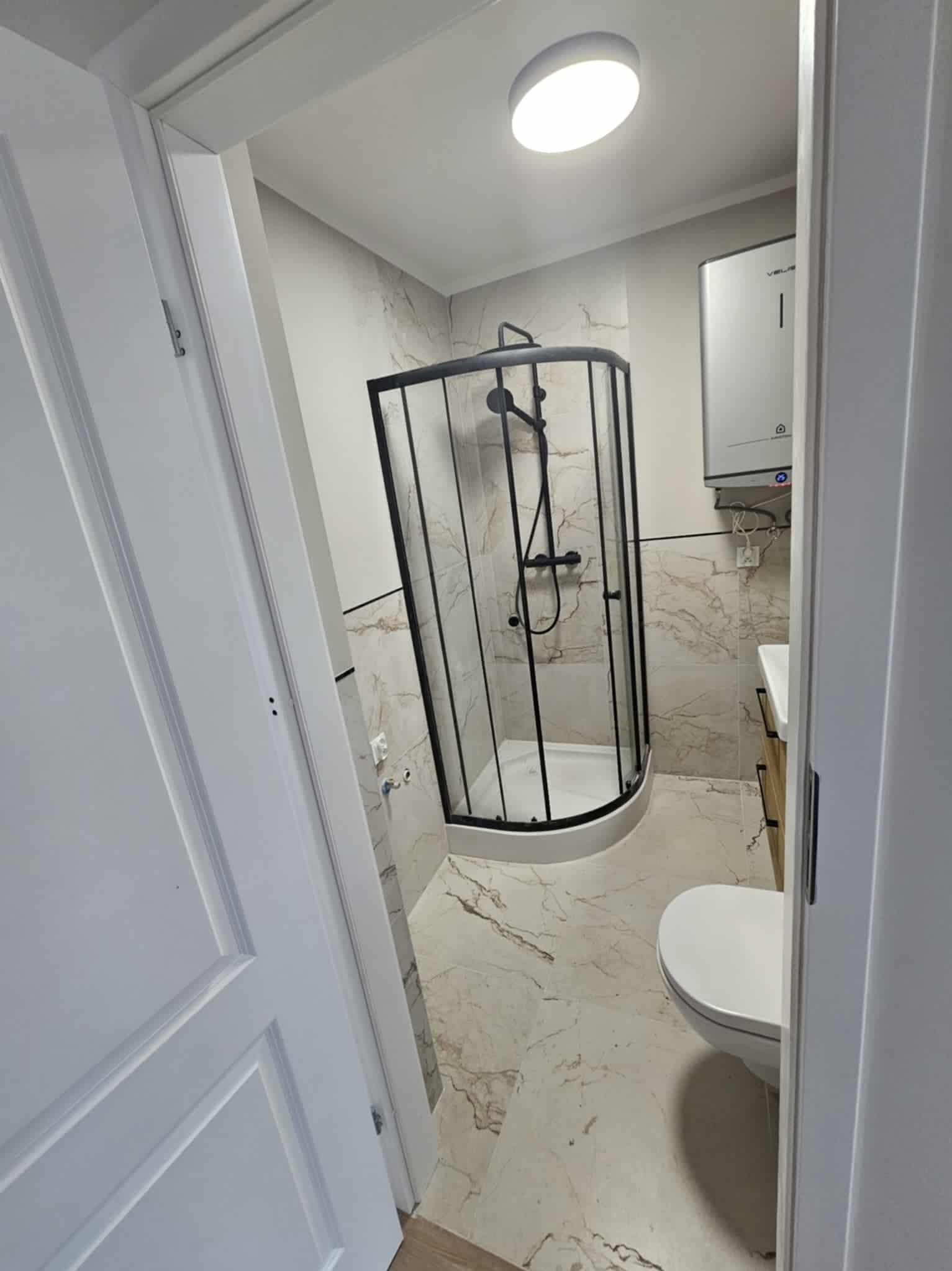 Wykończona łazienka z kabiną prysznicową z czarnymi profilami, marmurowymi płytkami i podwieszaną toaletą. Widok zza uchylonych białych drzwi.