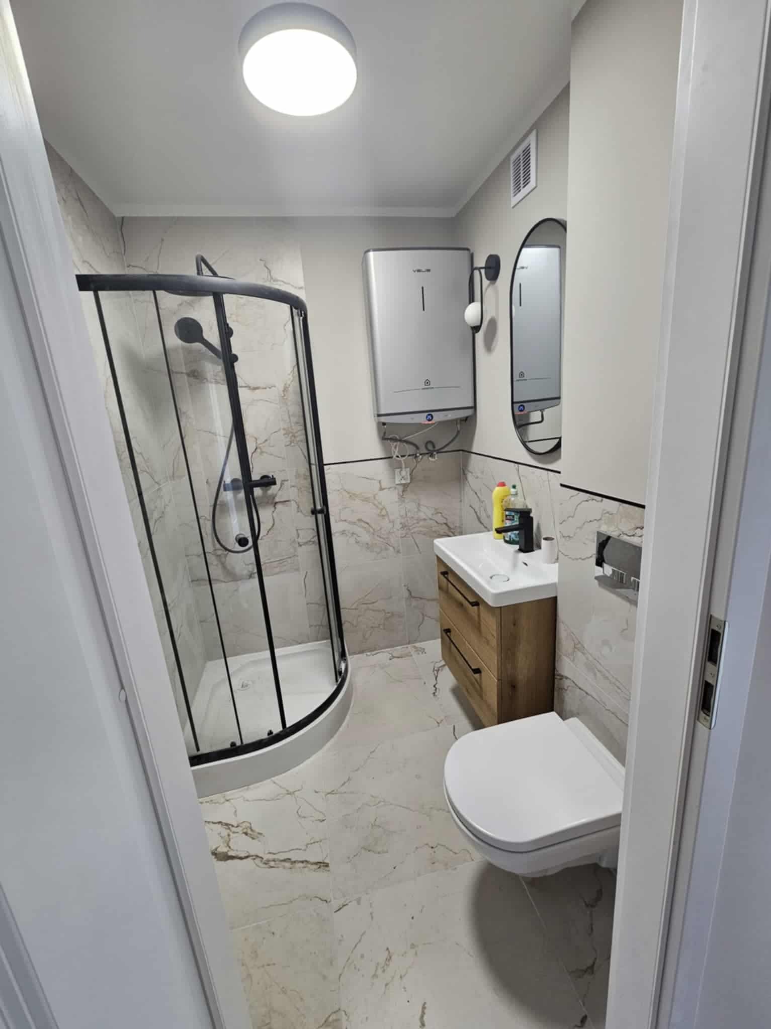 Wykończona łazienka z kabiną prysznicową typu walk-in z czarnymi ramami, umywalką z drewnianą szafką i marmurową podłogą. Widok zza uchylonych drzwi.