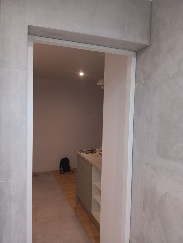 Wykończone przejście w łazience z szarymi płytkami, widok na wnętrze z szafką i torbą. Delikatne oświetlenie sufitowe. Minimalistyczny design.