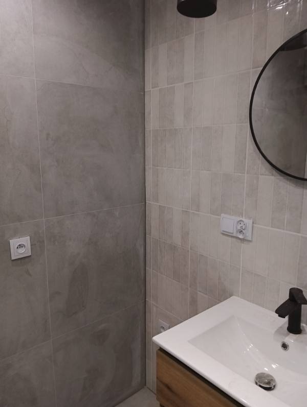 Wykończona łazienka z szarymi płytkami imitującymi kamień, okrągłym lustrem i umywalką nablatową z drewnianą szafką. Widoczne gniazdka elektryczne.