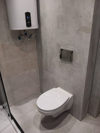 Wykończona łazienka z podwieszaną toaletą i grzejnikiem elektrycznym. Szare płytki imitujące beton na ścianach i podłodze. Minimalistyczny design.