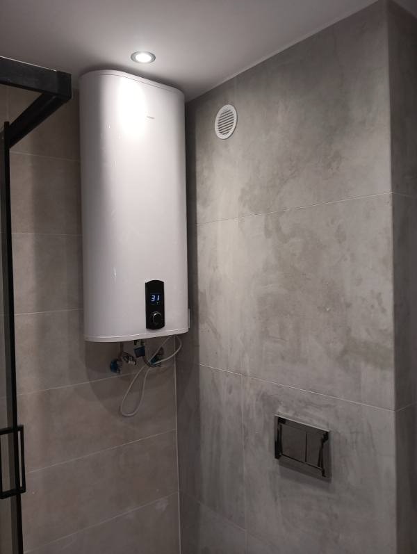 Wykończona łazienka z szarymi płytkami: biały bojler z wyświetlaczem, wentylacja i czarna rama kabiny prysznicowej. Minimalistyczny design.