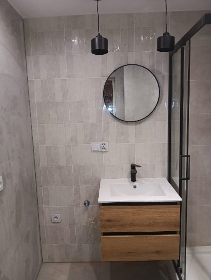 Wykończona łazienka z szarą, pionową glazurą, okrągłym lustrem, drewnianą szafką pod umywalką i czarnymi lampami wiszącymi.