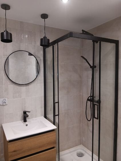 Nowoczesna łazienka z kabiną prysznicową w czarnej ramie, okrągłym lustrem i drewnianą szafką pod umywalką. Minimalistyczne lampy wiszące. Jasne płytki na ścianach.