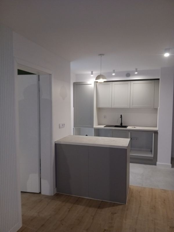 Nowoczesna kuchnia w odcieniach szarości i bieli. Widoczna wyspa kuchenna, szafki i oświetlenie punktowe. Podłoga drewniana, kontrastująca z chłodną kolorystyką pomieszczenia.