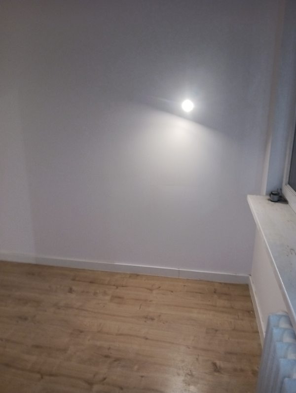 Wnętrze po remoncie: jasne ściany, drewnopodobna podłoga, listwy przypodłogowe. Widoczny fragment okna i grzejnika. Minimalistyczne oświetlenie punktowe na ścianie.