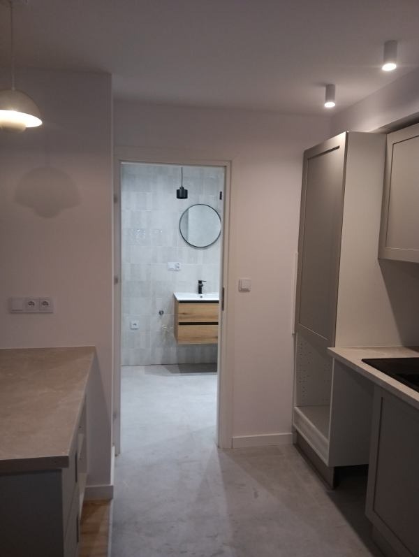 Nowoczesne wnętrze: szara kuchnia z widokiem na łazienkę z okrągłym lustrem i drewnianą szafką. Minimalistyczny design, neutralna kolorystyka, punktowe oświetlenie.