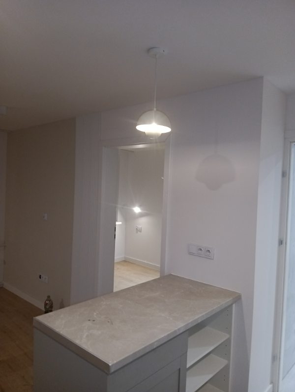 Wykończona kuchnia z wyspą o marmurowym blacie i białymi ścianami. Widoczna biała lampa wisząca i przejście do innego pomieszczenia. Nowoczesne gniazdka elektryczne.