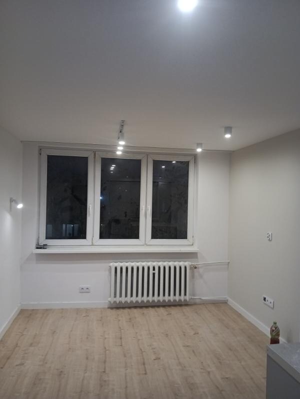 Jasne wnętrze po remoncie: panele podłogowe, białe ściany, okno z grzejnikiem. Minimalistyczne oświetlenie punktowe, gniazdka elektryczne i butelka w rogu.