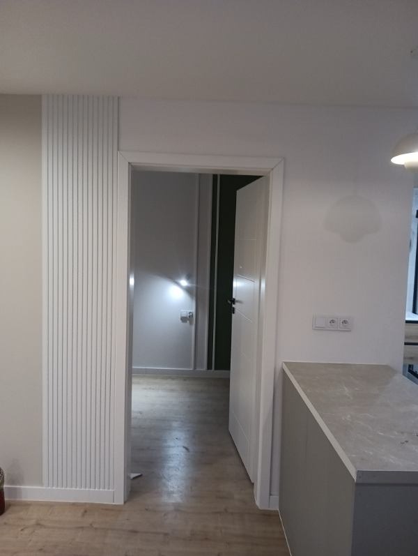 Białe panele ścienne dekoracyjne z MDF przy wejściu do pokoju. Widoczne otwarte drzwi i fragment blatu kuchennego. Minimalistyczny design wnętrza.
